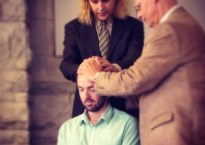 Michael is wearing a light green shirt, seated with eyes closed as two men in suits stand beside him with their hands on his head. Their eyes are closed and their heads are bowed.