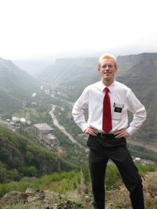 Isaac stands with his hands on his hips on a hill with a river valley behind him. He is wearing a white button down shirt with red tie, black pants, and an LDS missionary name tag.